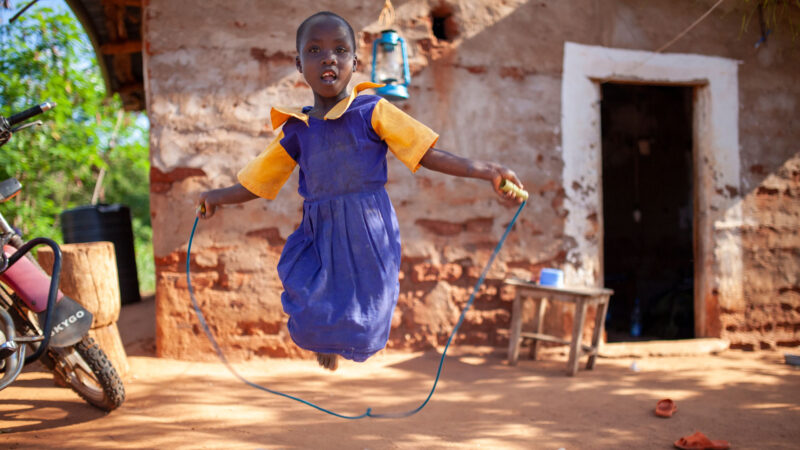 A child in a blue and yellow dress jumps rope outside a rustic building.