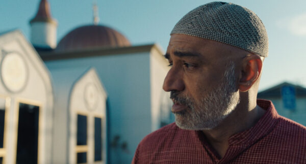 A man with a gray beard wearing a patterned cap stands outdoors in front of a building with a domed roof.