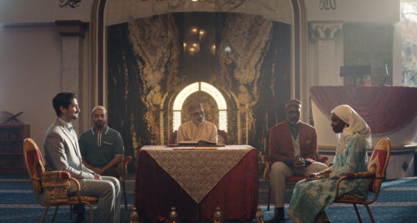 A group of five people sit together in an ornate room decorated with patterned walls and arches. The central figure sits behind a table covered with a lace cloth, with an open book in front, while the other four are seated in chairs facing each other in pairs. The room features luxurious chairs, patterned carpet, and elaborate wall designs, suggesting a formal or ceremonial gathering, possibly in a religious or cultural setting.