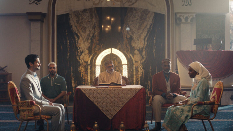 A group of five people sit together in an ornate room decorated with patterned walls and arches. The central figure sits behind a table covered with a lace cloth, with an open book in front, while the other four are seated in chairs facing each other in pairs. The room features luxurious chairs, patterned carpet, and elaborate wall designs, suggesting a formal or ceremonial gathering, possibly in a religious or cultural setting.