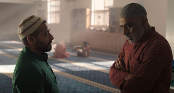 Two men wearing traditional caps and attire stand facing each other inside a spacious room with patterned blue carpet and sunlight streaming through large windows.