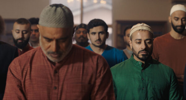 A group of men stand closely together in rows, heads bowed in solemn concentration, wearing traditional attire and prayer caps. The men at the front are dressed in red and green garments, with others filling the background, suggesting a communal or religious activity such as prayer in a mosque.