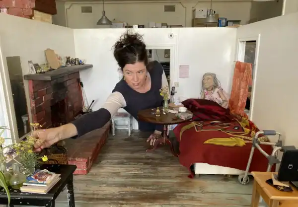 Woman arranging flowers in a miniature cozy room with a fireplace, a bed with a red blanket, and a doll sitting on the bed.