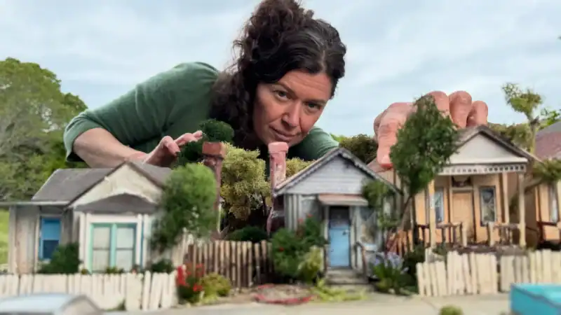 Woman with dark curly hair leaning over a miniature model of a neighborhood with small houses, trees, and cars, outdoors on a cloudy day.