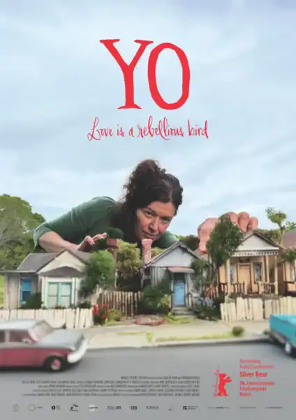 Close-up of a woman with dark curly hair leaning over a miniature model of a neighborhood with houses and trees, with a cloudy sky background, promoting the film 'Y.O' Love is a Rebellious Bird.