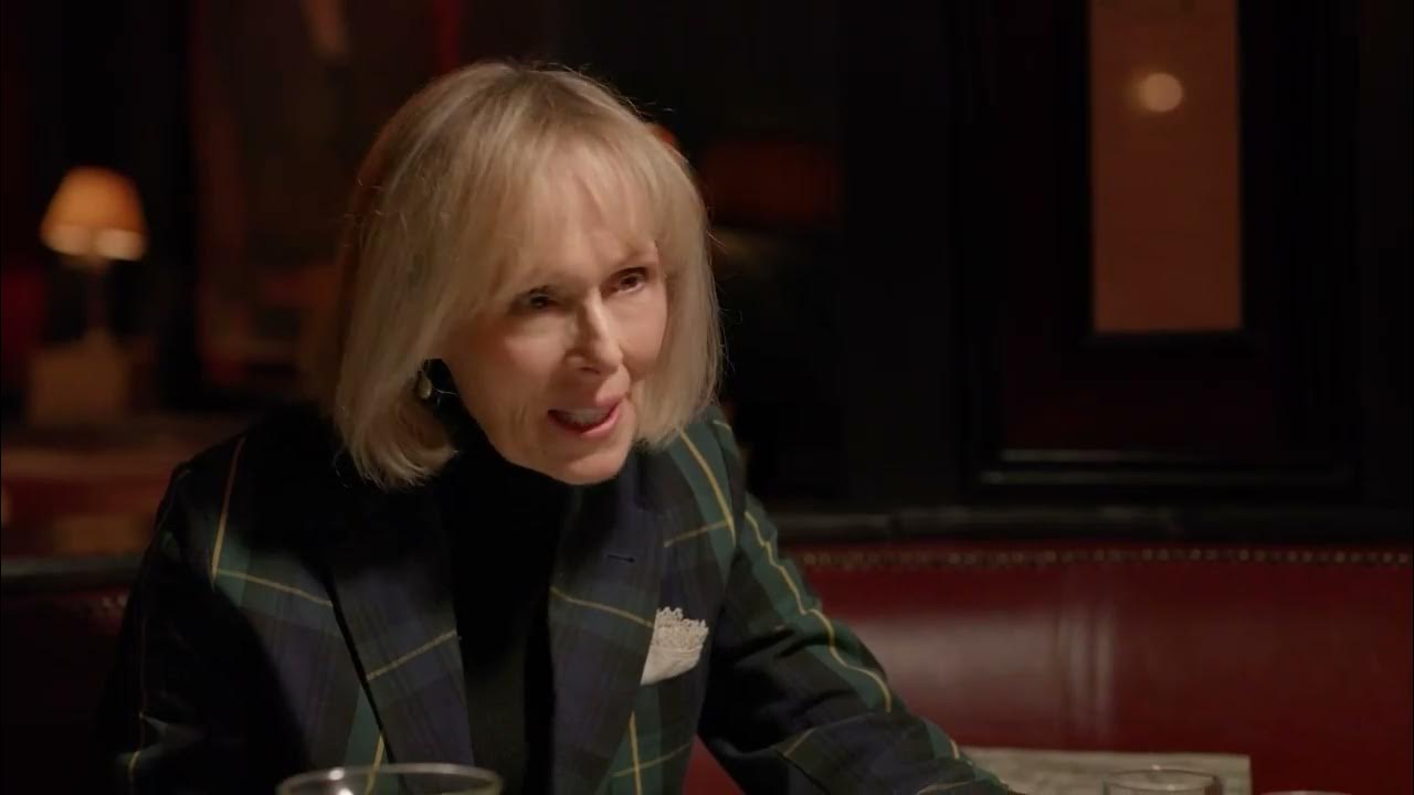 An woman with blonde hair and a plaid blazer sits at a table in a dimly lit space, speaking with a serious expression.