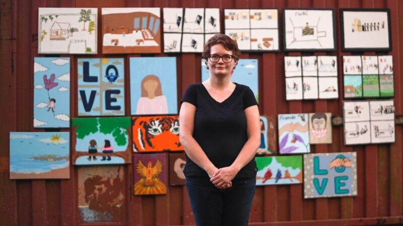 A woman with short hair and glasses stands in front of a wall displaying colorful paintings, including images of people, animals, and the word "LOVE."