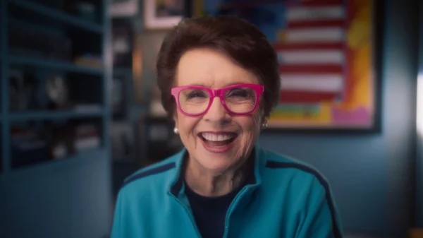 Woman with short brown hair and bright pink glasses smiles, wearing a blue zip-up jacket, seated indoors with an American flag artwork in the background.