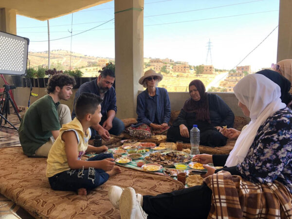 A group of people sits together on floor cushions around a large tray of food, enjoying a meal in a shaded outdoor area. The scene includes dishes of colorful salads, bread, dips, and drinks set on patterned mats, with views of hills and houses in the background. A film light and equipment are visible nearby, suggesting a casual, communal meal during a film shoot or gathering.