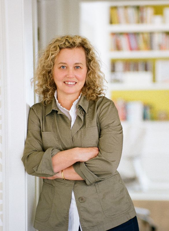 A woman with curly blonde hair, wearing a green jacket over a white shirt, stands indoors with arms crossed and smiles at the camera. A bookshelf is visible in the blurred background.