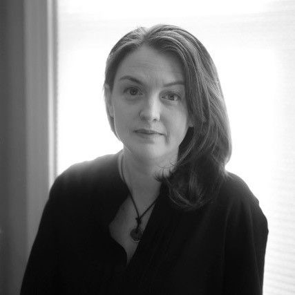 A woman with long dark hair, wearing a dark top and necklace, sits in front of a window with blinds. The image is in black and white.
