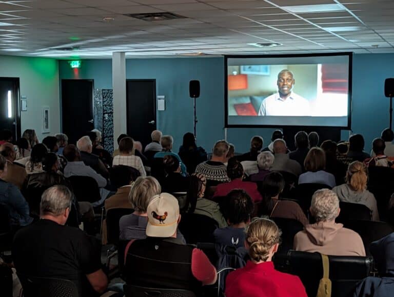A group of people sit in rows of chairs watching "I'm Carl Lewis!" on a large screen in a dimly lit room.