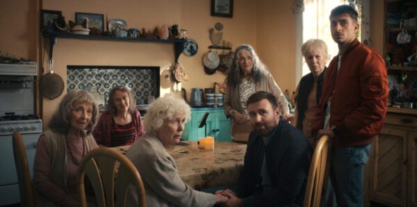 A group of seven people, diverse in age, gather around a kitchen table with a lit candle.