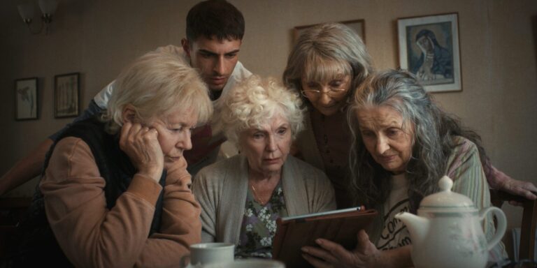 A group of elderly women and a young man are intently looking at a tablet in a cozy room.