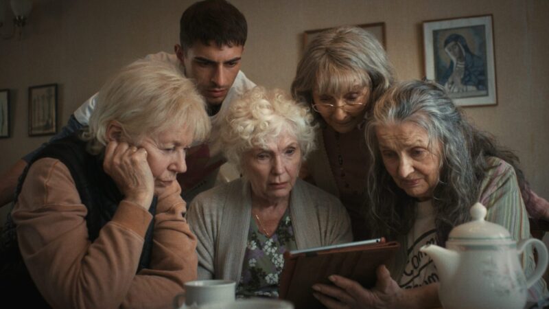 A group of elderly women and a young man are intently looking at a tablet in a cozy room.