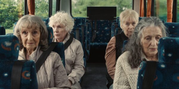 Four elderly women sit on a bus with patterned seats, gazing pensively outwards.