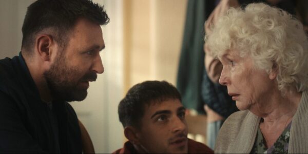 A serious scene with a bearded man on the left facing an elderly woman with curly white hair on the right.