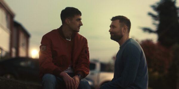 Two men sit facing each other on a stone surface outdoors. One wears a red jacket, the other a blue shirt.