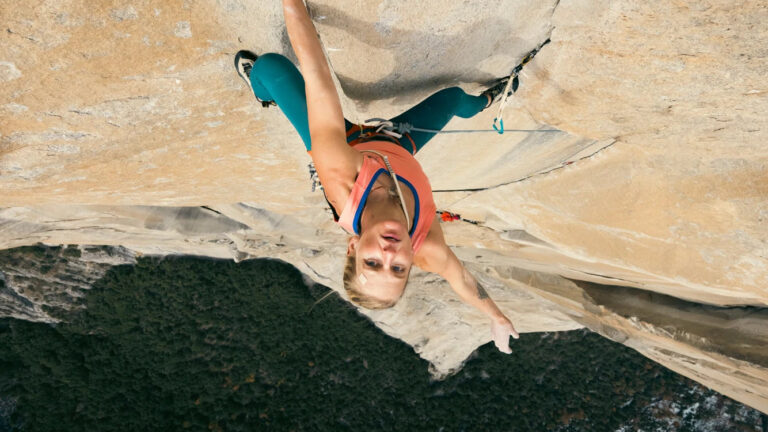 A person wearing climbing gear ascends a steep rock face, gripping the rock with one hand and looking upward toward the camera. Dense forest is visible far below.