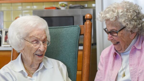 Two elderly women with white hair sit indoors, smiling and talking with each other. One wears glasses and a pink shirt, the other wears a white blouse.