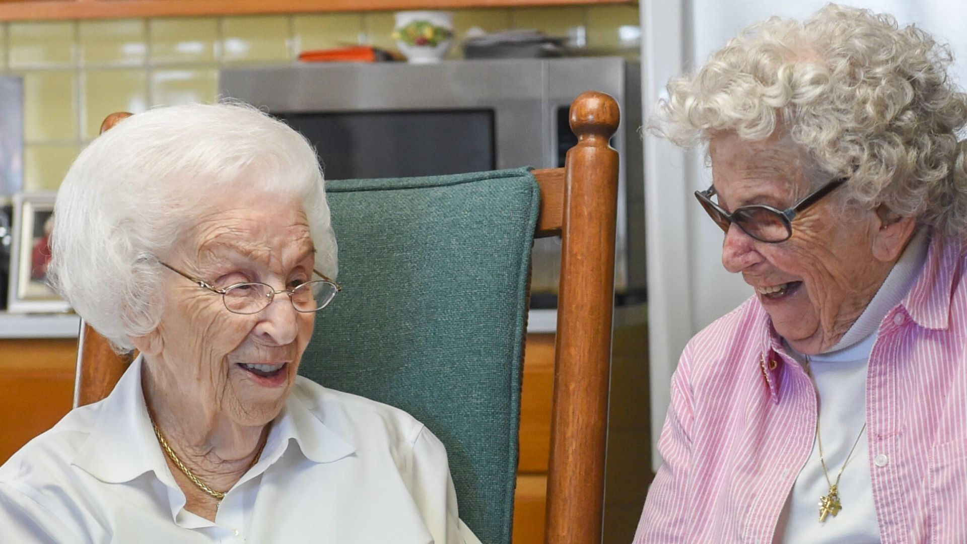Two elderly women with white hair sit indoors, smiling and talking with each other. One wears glasses and a pink shirt, the other wears a white blouse.