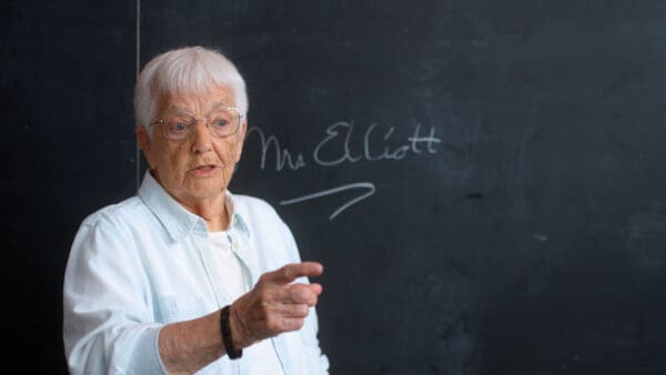Jane Elliott teaching in front of a blackboard with her signature visible.