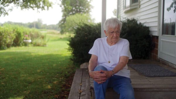 Elderly woman with white hair and glasses sitting on a porch, with a lush green garden in the background.