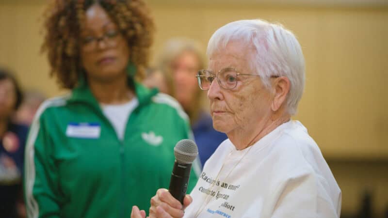 A close-up of an older woman, holding a microphone, giving a speech to a group of people.