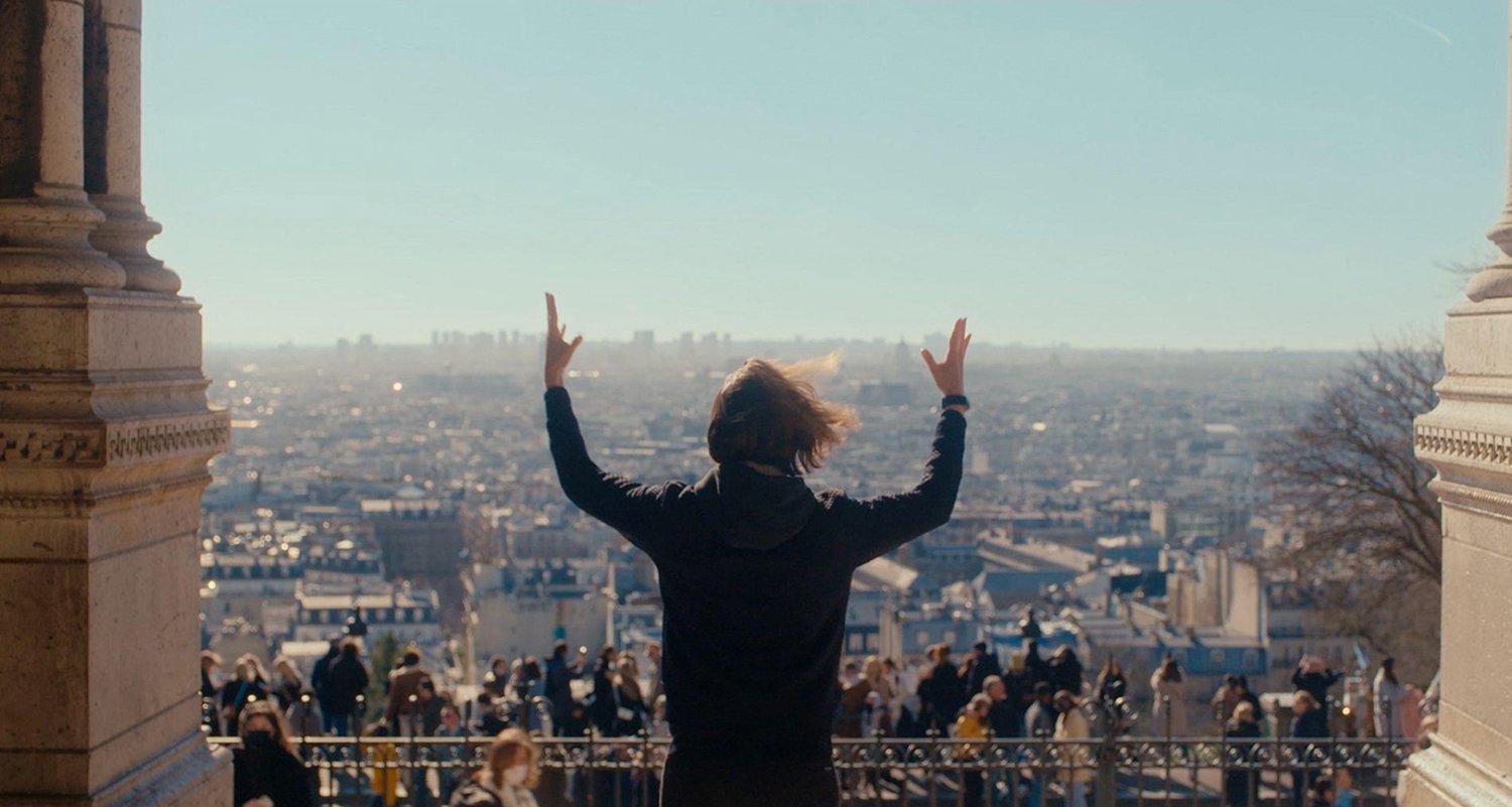 A person stands with raised arms facing a cityscape, overlooking a crowded urban area from a high vantage point on a sunny day.