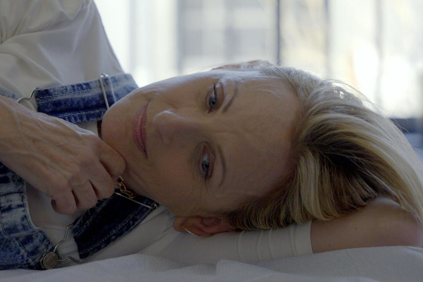 A woman with blonde hair lies on her side, resting her head on her arm and looking forward thoughtfully, wearing a white shirt and denim overalls.
