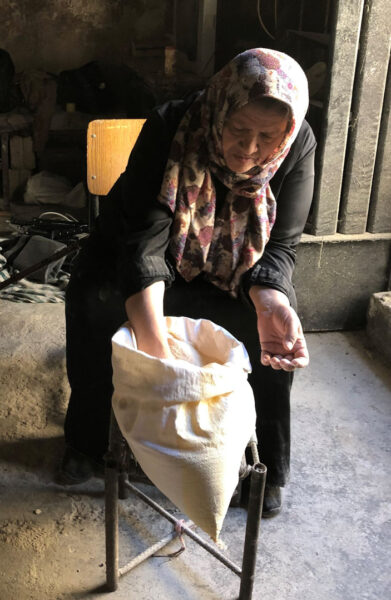 A person sits in a rustic, dimly lit space, wearing a floral-patterned headscarf and dark clothing. They are focused on milling or sifting flour, reaching into a white cloth bag placed on a stand in front of them. The scene conveys a sense of concentrated manual labor in a traditional or rural setting.