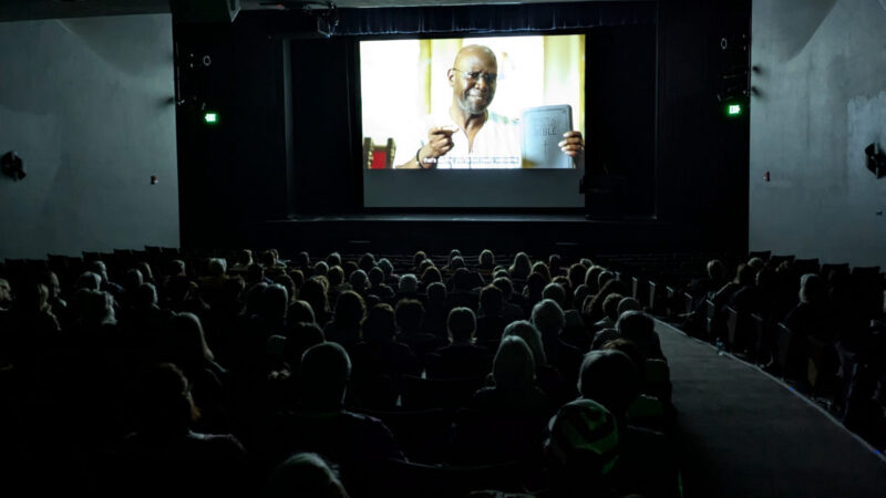 Audience seated in a dark movie theater watching a film; the screen shows a man holding up a Bible and speaking, with subtitles visible.