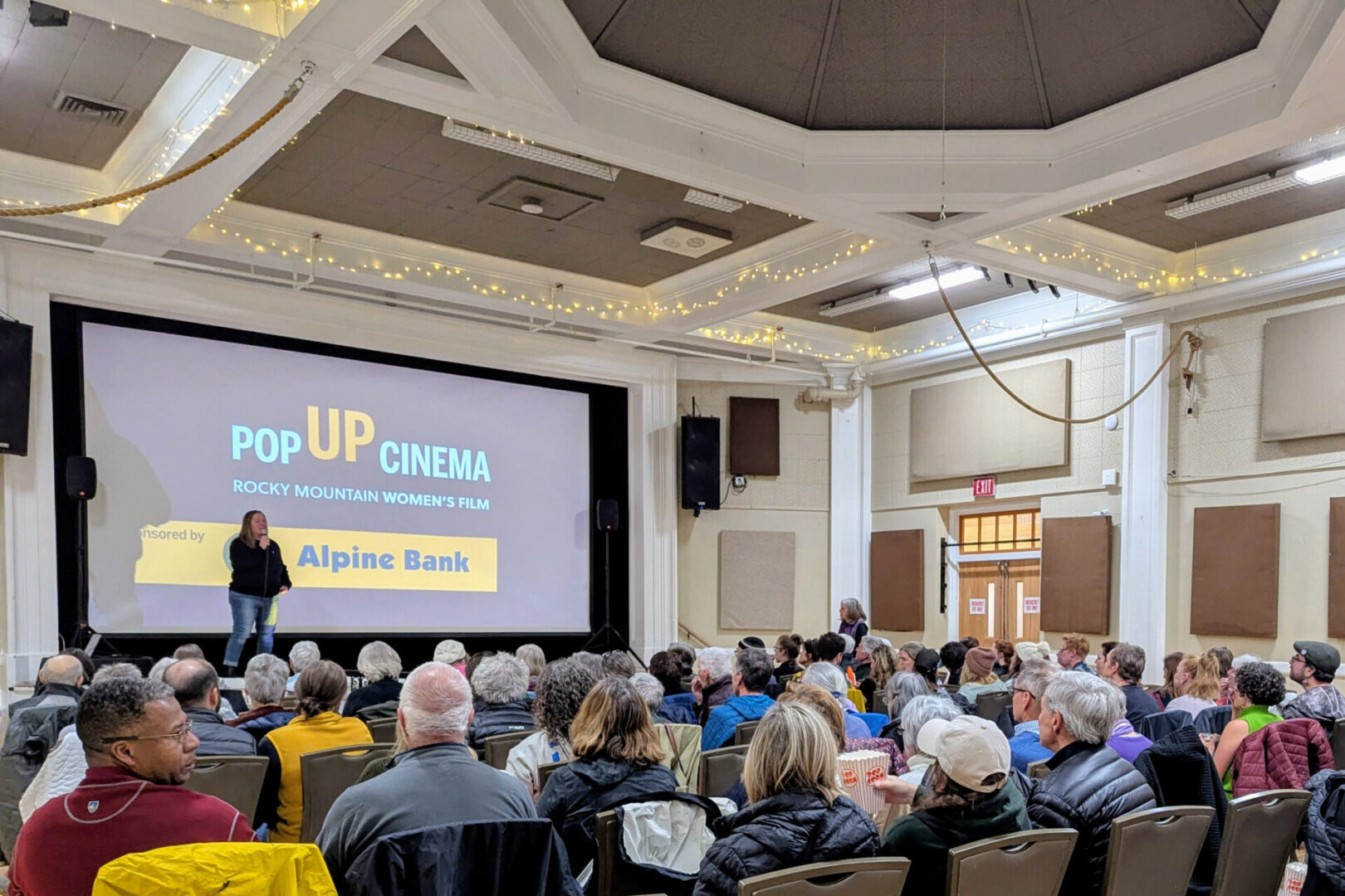 Audience seated in a hall watches a presentation on a large screen that reads "Pop Up Cinema Rocky Mountain Women's Film" with Alpine Bank logo, under string lights.