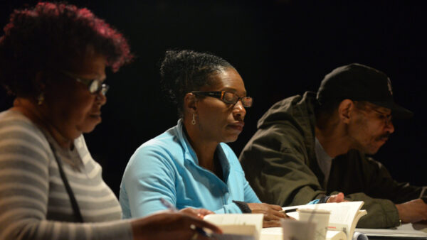 Three adults sit at a table reading books and taking notes, focused on their work under bright lighting.