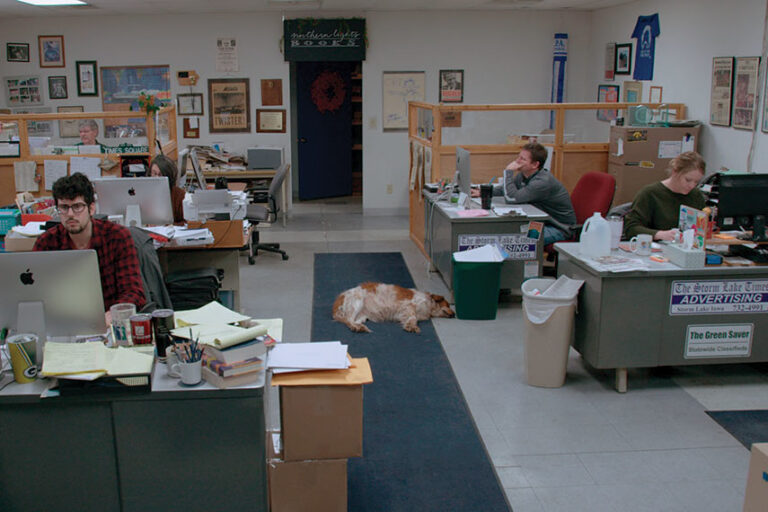 An office with several people working at desks, papers and computers on surfaces, and a dog lying on the floor in the center aisle.