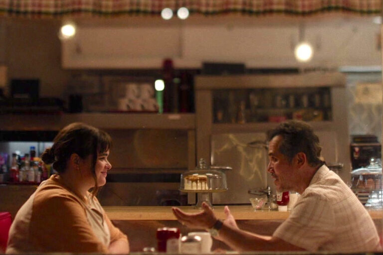 Two people sit across from each other at a diner counter, engaged in conversation.