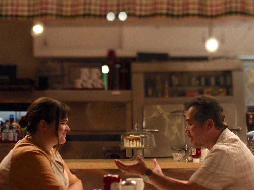 Two people sit across from each other at a diner counter, engaged in conversation.