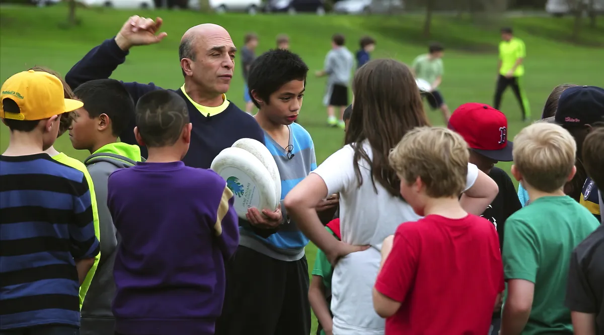 An adult coach instructs a group of children outdoors on a grassy field, holding two frisbees while the kids listen attentively.