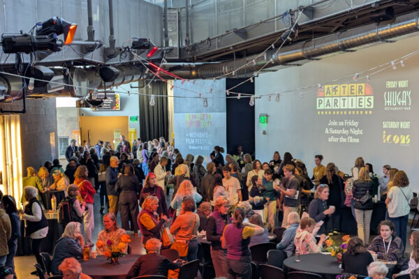 A large group of people gathers indoors at a venue with tables, flowers, and event signs on the wall promoting after parties and film festival activities.