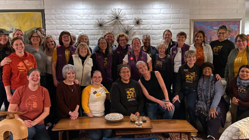Large group of people, many wearing festival shirts or vests, gathered together indoors on couches and chairs, smiling for a group photo at a casual social event.