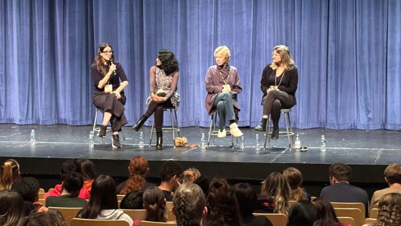 Four filmmakers sit on chairs speaking on a stage in front of a blue curtain, with an audience of high school students seated and facing them.